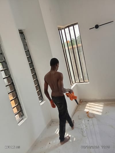 The image shows a young man, shirtless and wearing dark pants and orange gloves, operating an orange blower in what appears to be an interior construction or renovation site. He is standing and facing away from the camera, directing the blower towards the floor. The room has white walls and a tiled floor with debris scattered around. There are two narrow windows with metal bars on the left wall, and a larger window with similar bars on the right wall, overlooking an outdoor scene with greenery. The lighting suggests it is daytime. The bottom left corner of the image displays "REDMI A3" and the bottom right corner shows the date and time "06/18/2026 11:09". This scene is located in Nawfia, Nigeria.