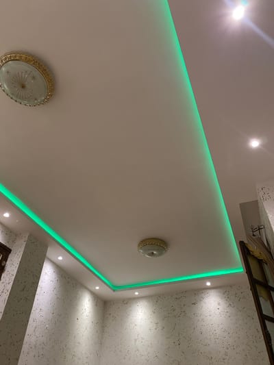 The image shows a view of a ceiling with recessed lighting and decorative elements. The ceiling is white and features a green LED strip light installed along the perimeter of a recessed section. There are two flush mount ceiling light fixtures visible, one in the upper left and another in the lower center of the image. Both fixtures have a gold-colored trim and a decorative glass cover. The walls appear to be textured with a light color and a speckled pattern. In the upper right corner, a bright light source is visible, possibly an overhead lamp. The image captures an interior scene, likely a residential or commercial space, in Baraki, Algeria. No people are visible in the image.