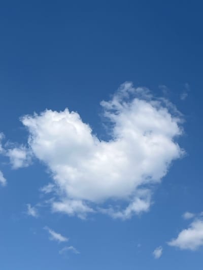 The image shows a bright blue sky with a large, puffy white cloud in the center-right of the frame. There are also a few smaller, wispier clouds scattered around. The location is not identifiable in the image. No people, subjects, objects, actions, or interactions are visible other than the clouds and the sky.