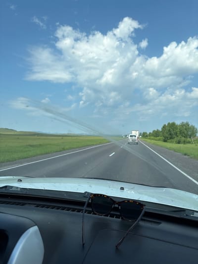 The image depicts a view from inside a vehicle looking out at a highway. The foreground shows a car's dashboard with a pair of sunglasses resting on it. The road stretches into the distance, with other vehicles, including a white SUV and a truck, visible on it. The landscape on either side of the road consists of green fields and trees under a blue sky with white clouds. The scene suggests a journey on a sunny day in an open, rural area. Based on the image alone, the specific location in Borodino, Russia cannot be determined.