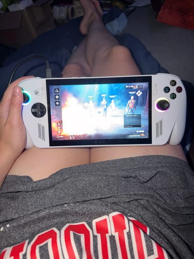The image shows a person holding a white handheld gaming console, likely an ASUS ROG Ally, with a game displayed on its screen. The game appears to be Dead by Daylight, with four characters visible on the screen, two survivors and two killers in a dimly lit, wooded environment. The console has a white exterior with black buttons and joysticks, and the screen is illuminated with bright colors from the game. The person's hands are visible holding the device, with the left hand on the left joystick and the right hand on the right joystick and face buttons. The person is wearing a grey t-shirt with red lettering, and is sitting on a blue surface, possibly a couch or bed. The background is blurred, but suggests an indoor setting with some boxes and furniture visible. The lighting is low, with the primary light source coming from the console's screen and RGB lighting around the joysticks. There is no clear indication of the specific location within Portland, United States of America, as the image focuses on the person and the gaming device.