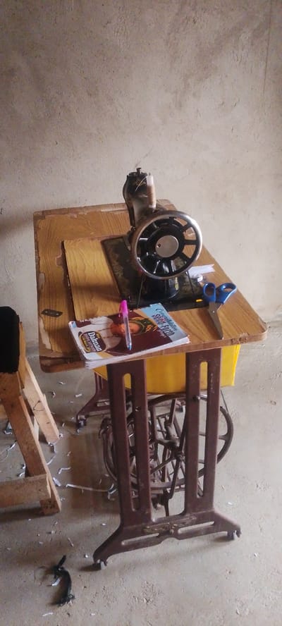 The image depicts an antique sewing machine situated on a wooden table in Doka-Tofa, Nigeria. The machine itself is dark metal with a large wheel. Resting on the table next to the sewing machine are a pink pen, a book with the title "Congratulations Send-Forth" visible, and a pair of blue scissors. The table is supported by a dark metal stand with intricate scrollwork. In the background, a plain, textured wall dominates the scene. To the left of the table, a portion of a wooden structure is visible, and white paper scraps are scattered on the concrete floor around the sewing machine. A dark, metallic object, possibly a detached handle or foot, lies on the floor to the lower left of the frame. The overall environment suggests a functional space, possibly a home or workshop, where sewing activities take place.