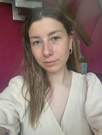 A woman with light skin, brown hair, and gray eyes is positioned at an oblique angle in the image. Her face features visible freckles, particularly on her nose and cheeks. She is wearing a light-colored shirt with a textured pattern. A silver hoop earring is visible in her left ear. The background consists of a red wall, a wooden object, and what appears to be a stainless steel structure. There is no indication that the photo was taken in the city of Чёрное, Russia.
