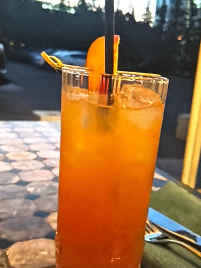The media file shows a close-up, horizontally oriented shot of a drink in a tall glass. The drink is a vibrant orange color and appears to be a cocktail or juice, possibly with ice. A black straw is visible inserted into the drink, along with a slice of orange or lemon as a garnish, attached with a thin yellow loop. The glass itself has a textured rim.
The background is dark and somewhat blurred, suggesting an indoor setting, possibly a bar or restaurant. Hints of a patterned surface, possibly a tablecloth or placemat, are visible at the bottom of the frame, with a circular, mosaic-like design in muted blues and grays. A portion of a green cloth or garment is also visible in the upper right corner, with a blue and white striped ribbon or fabric draped over it.
The lighting suggests it might be evening or indoors with artificial lighting. No people are clearly visible, and no text is discernible. The overall impression is a still life of a refreshing beverage.