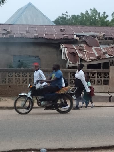 The image shows a street scene in what is likely Biliri, Nigeria. A man is riding a black motorcycle on a paved road. A woman in a white shirt and black skirt walks alongside a child with a red backpack. Another woman in a white shirt walks nearby, with a woman with a red headscarf standing behind them. In the background is a building with a crumbling rusty metal roof, a concrete wall, and a pyramidal structure on the roof. The environment appears to be a somewhat impoverished urban or peri-urban area.
