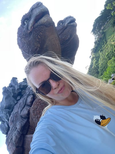 In the city of Thôn 1, Vietnam, a young Caucasian woman with long blonde hair and sunglasses takes a selfie. She is wearing a light blue t-shirt with a cartoon duck graphic on the left breast. Behind her, a large, textured rock formation is visible, and two large, dinosaur-like figures with scaly skin and open mouths are positioned above and behind her. Lush green foliage and trees are present on a hillside to the right of the frame, and the sky is bright and overcast.