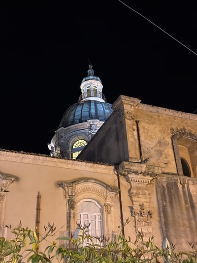 Here's a description of the image:
The image, taken at night in Ragusa, Italy, features a building with an ornate dome, likely a church or cathedral, sharply contrasted against a black sky. The architecture of the building is visible with its stonework, archways, and decorative detailing. A tree with visible foliage partially obstructs the view of the building. The dome is lit, suggesting the presence of artificial lighting. A wire runs across the dark sky. The building's facade shows architectural details and variations in texture, indicating age.