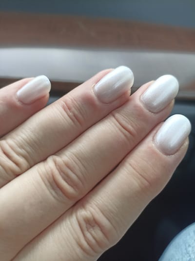 A close-up, overhead shot captures a person's hand with five fingers, all adorned with milky white, pearlescent nail polish. The nails are neatly shaped and have a smooth, almost satin finish. The hand is positioned with fingers slightly curved, showcasing the texture of the skin and the cuticles around the nails. The background is softly blurred, suggesting an indoor setting with diffused lighting. To the top of the image, there's a hint of a wooden surface and a light-colored stripe, possibly part of a desk or furniture. The lower portion of the image is dark, with a slight blueish tint and a textured pattern, perhaps denim fabric. No text is visible in the image.