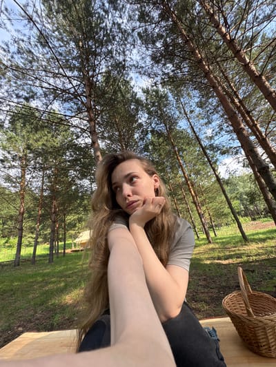 A young woman with long, light brown hair is lying down outdoors, with her head propped on her hands. She is wearing a light gray t-shirt and dark pants. Her expression is relaxed and contemplative. Behind her, a woven basket sits on a wooden surface. The setting is a wooded area with tall pine trees and a grassy field. The sun is shining, creating dappled light and shadows on the ground. The scene appears to be taking place on a clear day. The overall mood is peaceful and serene, suggesting a moment of quiet enjoyment in nature. The location context provided is "Гольяны, Russia". There is no visible text in the image.