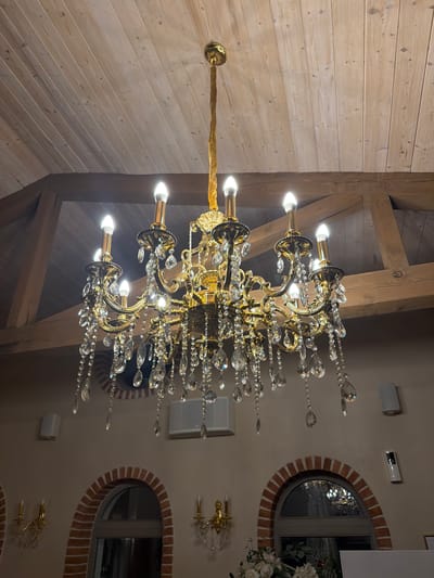 This is a medium shot, eye-level, indoor photograph of a grand, ornate, gold-colored chandelier with numerous crystal accents hanging from a wooden planked ceiling. The chandelier has multiple arms, each holding a light bulb that is lit, and is adorned with teardrop-shaped crystals that refract light. The ceiling features exposed wooden beams, adding to the rustic yet elegant ambiance. Below the chandelier, the wall is painted a neutral tone and features two arched windows with brick trim. Several wall-mounted speakers and what appears to be a security camera are visible on the wall. In the foreground, part of a floral arrangement is visible. The overall setting suggests an interior space such as a restaurant, banquet hall, or a well-decorated home, possibly in Kocherinovo, Bulgaria. There are no people visible in the image.