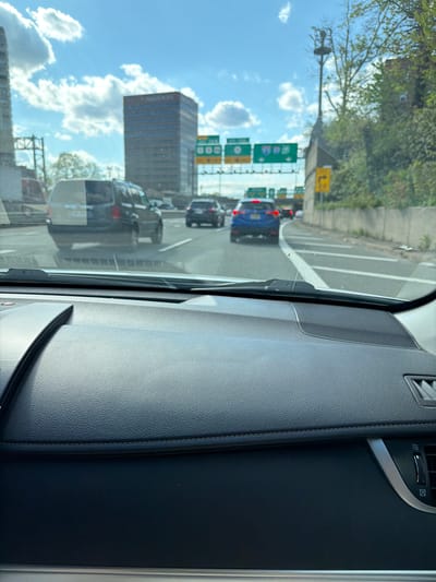 The image, taken from the interior of a vehicle, shows a traffic scene on a highway in Fort Lee, New Jersey, United States of America. The highway appears congested, with multiple vehicles visible, including a blue sedan and a dark-colored van. Signage indicates highway exits and directions. A tall office building is visible in the background. The sky is blue with scattered clouds. The vehicle's dashboard and interior panels are prominently featured in the foreground.