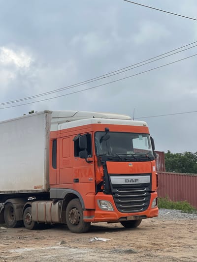A DAF truck with an orange cab and a white trailer is parked on a dirt road in Karmajiji, Nigeria. The truck has the number "680" on its side, along with the logo "SIRA". The trailer has text that reads "TRANS VERHUIZINGEN" and "VERGOEDEL OPSLAG". There are shipping containers in the background, and trees and foliage are visible. The sky is overcast. The front of the truck appears to have some damage.