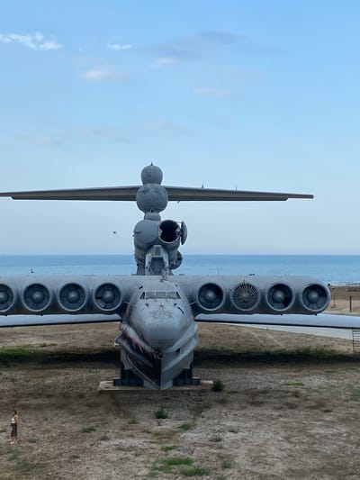 A static, wide-angle, eye-level shot captures the front of a large, gray, military aircraft with its nose pointed towards the viewer. The aircraft is positioned on a dry, dusty ground with sparse vegetation. The ocean stretches out in the background, meeting a hazy sky with scattered clouds. A small, lone figure of a woman in a white top and dark skirt stands to the left of the aircraft, facing away from the camera. The aircraft features multiple engines mounted on its wings, and its front is decorated with a painted shark mouth graphic. The scene appears to be a museum or a preserved monument, given the aircraft's static display and the surrounding environment.