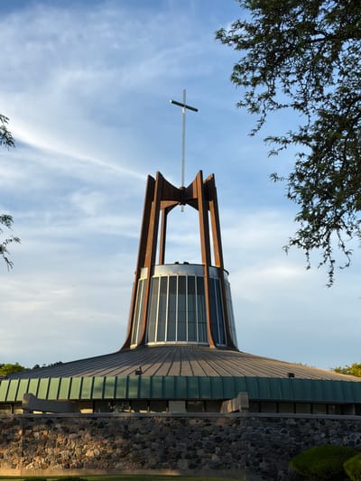 The image depicts a low-angle view of the Prince of Peace Parish church building in Niles, Illinois, USA. The building features a modern architectural style with a distinctive cross atop a brown wooden framework. The church incorporates a cylindrical structure with windows and a sloping roof with a stone exterior wall. The sky behind the building is blue with streaks of white clouds. Tree branches and foliage are partially visible in the upper and lower portions of the image.