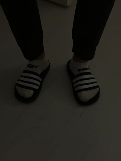 The image shows a close-up view of a person's feet wearing white socks with black stripes and dark blue slides. The person is wearing black pants. The floor appears to be a light-colored laminate. The scene is dimly lit. There is no information available to determine the location in the city of Balbriggan, Ireland.