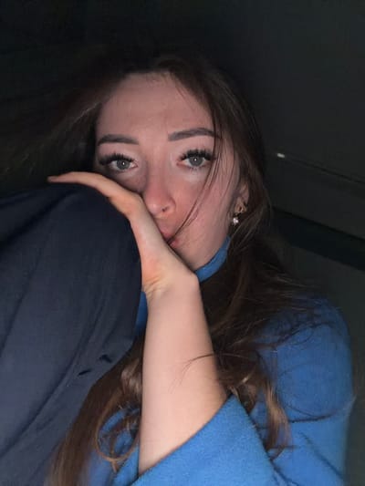 This is a close-up, low-angle, indoor photograph of a young woman. She has long, brown hair that is falling around her face. She is wearing a blue hooded sweatshirt and dark pants. Her face is partially obscured as she rests her head on her arm and knees. Her eyes are open and looking towards the camera, with long eyelashes. She is wearing a small, sparkling stud earring in her visible ear. The lighting is dim, suggesting it might be evening or a poorly lit indoor space. The woman's expression appears thoughtful or perhaps a little melancholic. The background is dark and indistinct, offering no specific clues about the location beyond the fact that it is indoors. The context provided places the image in Yarykovo, Russia. There is no visible text in the image.