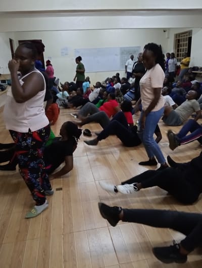 This image depicts a lively indoor group activity, likely a fitness or exercise session, held in what appears to be a community hall or spacious room in Riara Ridge, Kenya. The setting features light-colored walls and a warm-toned wooden or tiled floor.
The room is filled with numerous individuals, predominantly adults, who are engaged in various physical movements. In the foreground on the left, a woman wearing a light pink sleeveless top and dark patterned trousers stands, her hand near her face as she looks downwards, appearing contemplative or focused. Slightly to her right, another woman in a light pink t-shirt and blue jeans stands with her back partially to the camera, observing the activities.
The majority of the participants are seated or lying on the floor, actively performing exercises such as leg raises or crunches, indicated by their angled bodies and extended legs. Their diverse attire suggests a casual, athletic environment. A large group fills the mid-ground and background, with most individuals similarly positioned on the floor, although a few can be seen standing near the back.
On the back wall, a white board or screen is visible, containing some faint, illegible writing. Windows are also present on the right side of the background. The indoor space is well-lit, providing clear visibility of all participants. The overall scene conveys a sense of group participation and physical exertion.