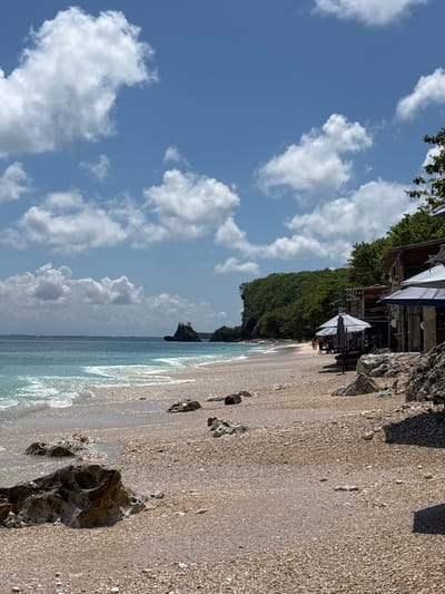 The image depicts a beach scene in Uluwatu, Indonesia. The sky is blue with scattered white clouds. The beach is made of light-colored sand and pebbles, with some scattered dark rocks near the shoreline. The ocean is a light blue-green color, with small waves lapping at the shore. Along the edge of the beach, there is a small restaurant named “SUKON HERE” with parasols and beach chairs. Dense green vegetation covers a cliff or hillside behind the restaurant. There are a few people visible under the umbrellas.