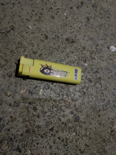 A single yellow (or lime green) lighter is centrally placed on a dark, textured surface, likely concrete or asphalt, exhibiting small embedded stones and general wear. The lighter has a graphic design featuring the word "BILLIARDS" above an 8-ball and a crown-like symbol. On another side, the word "ZABATA" is visible, oriented upside down relative to the main graphic. A barcode with the numbers "979131 042099" is printed near the top of the lighter. The lighting appears dim, suggesting artificial light or low natural light. There are no people or other objects in the frame, and no specific event is taking place. The overall impression is of an object dropped or resting on a ground surface, possibly in Jama A, Nigeria, as provided in the context.