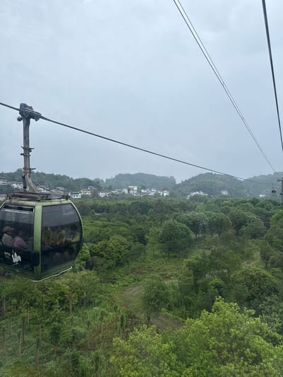 The image shows a green cable car ascending a mountain in Yongding District, China. Passengers are visible inside the car. Lush green vegetation covers the mountain, with scattered white buildings visible on the slopes. Overhead cables extend from the cable car towards a higher station. The sky is overcast and gray.