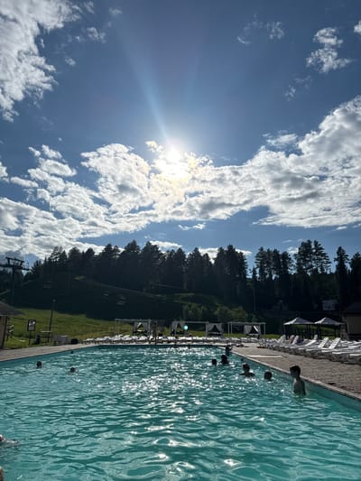 The image depicts an outdoor swimming pool filled with blue, rippling water, reflecting the bright sunlight. Several people are visible swimming or wading in the pool, some with their heads above water. Along the edge of the pool, lounge chairs and white canopies are arranged on a wooden deck. In the background, a lush green hillside is covered with a dense forest of pine trees. A ski lift with chairs is also present on the hillside, suggesting the location is likely a recreational area. The sky is a vibrant blue, dotted with fluffy white clouds and a bright sun with visible rays. The scene appears to be in Nechkino, Russia.