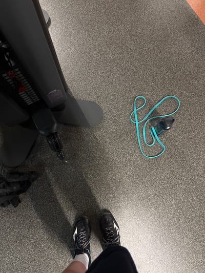 A low-angle, eye-level shot captures a gym setting in Enschede, Netherlands. In the foreground, two black and white athletic shoes are visible, worn by an unseen individual. The shoes are placed on a grey speckled carpeted floor. To the left, a black exercise machine, possibly a weight machine, dominates the frame, with its base resting on the same carpet. To the right of the machine, a teal resistance band is loosely coiled on the floor, with a black water bottle nestled within its loops. The lighting suggests an indoor environment, likely a gym, with shadows cast by the exercise machine.