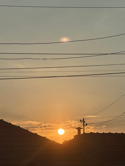 The image depicts a sunset in Dalgopol, Bulgaria, with the sun low on the horizon, casting a warm, golden glow. The scene is dominated by a silhouette of a dark, textured surface, possibly a building or a hillside, that takes up the right side of the frame. A prominent utility pole with wires is visible against the sky, adding an element of human infrastructure to the natural landscape. The sky is a soft gradient of pale blue to warm orange, with scattered clouds tinged by the setting sun. Several dark electrical wires stretch horizontally across the frame, creating a strong graphic element that divides the sky and adds depth to the composition. There are no people visible in the image. The overall mood is serene and peaceful, characteristic of a quiet evening. The time of day is clearly sunset.