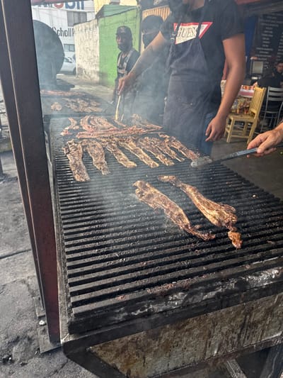 The image depicts an outdoor street food scene in Nezahualcóyotl, Mexico, centered around a large metal grill. Numerous strips of meat are cooking on the blackened grates, emitting significant smoke.
In the foreground and midground, a male individual wearing a black "GUESS" t-shirt and a dark apron is actively turning pieces of meat on the grill using metal tongs. Tattoos are visible on his right forearm. To his right, a partially visible hand and forearm hold a long-handled metal spatula, also interacting with the meat. Further left and behind the primary cook, another male individual wearing a black cap and a dark shirt, possibly with a face covering, stands near the grill.
The grill itself is constructed from heavy-gauge dark metal, with multiple horizontal grates spanning its surface. Its lower structure shows signs of extensive use and charring. The ground surrounding the grill is a dark, uneven paved surface.
In the background, behind the grill, there is a street or alleyway. A white building with a partially visible "Mobil P Scholl" sign and a green-painted wall are present. To the right, a dark menu board with white text lists various meat cuts and prices in Spanish, such as "COSTILLA ARGENTINA" and "NEW YORK." Several patrons are seated at dark tables with a yellow wooden chair visible, suggesting a casual dining area. The overall lighting suggests daylight, possibly an overcast day or late afternoon.