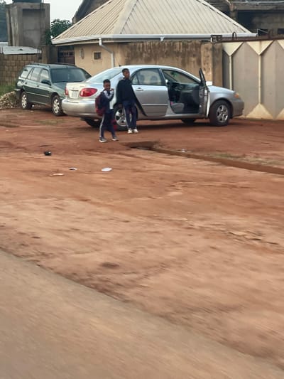 In Barakin Sabawa, Nigeria, a silver sedan with its driver's side door open is parked on a dirt road. Beside it, a dark green SUV is also parked. Three young people are near the sedan. Two of them are standing close together, one with their arm around the other, and appear to be wearing matching dark blue tracksuits with red accents. The third person, wearing a dark jacket, is standing behind them. The ground is a reddish-brown dirt, and a building with a corrugated metal roof is visible in the background. The scene suggests a casual, everyday moment. The lighting indicates it is daytime, likely overcast or late afternoon, given the soft shadows.