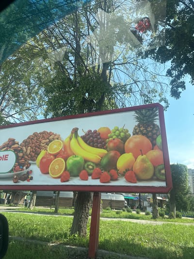 The image is a photograph taken from inside a vehicle, looking out at a billboard advertising a variety of fruits, nuts, and candies. The billboard is mounted on a red metal frame and is supported by red poles. The artwork on the billboard features a diverse arrangement of fruits, including oranges, apples, bananas, grapes, strawberries, pears, kiwis, and a pineapple. To the left, there are piles of nuts and a section of assorted cookies and candies. The billboard is positioned outdoors, with trees and a bright blue sky visible in the background. The lower portion of the image shows a grassy area and the side of a street or road, with some buildings and trees in the distance. A glimpse of urban infrastructure, such as what appears to be a market or small shops, can be seen behind the billboard. The overall scene suggests a commercial area or roadside advertising in an urban environment. The reflection of trees and a blue sky is visible on the vehicle's windshield, indicating the viewpoint. Without specific landmarks or clear signage in the background, pinpointing the exact location within Magnitogorsk, Russia, is not possible based solely on the visual information. However, the presence of a billboard, trees, and buildings is consistent with a typical street scene in a Russian city.