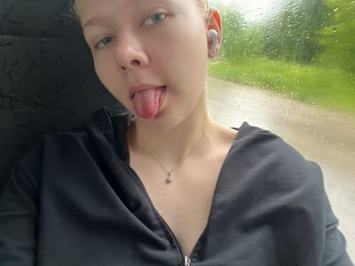The image shows a close-up, upside-down selfie of a young person. The subject is wearing a black zip-up hoodie, with a silver necklace featuring a small, clear pendant resting on their chest. They have blue eyes and blonde hair, and are wearing white wireless earbuds in their left ear. Their tongue is sticking out, and their expression appears playful or mischievous.
The background suggests an indoor setting looking out a window. The window pane is covered in raindrops, indicating that it is raining outside. Through the rain-streaked glass, a blurred green landscape with trees is visible, hinting at a daytime setting. The overall mood is casual and perhaps a bit whimsical, given the subject's playful pose.
The photograph appears to be taken in Krasnokamsk, Russia. There is no visible text in the image.