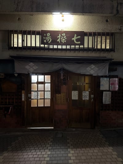 The image depicts the exterior of a traditional Japanese bathhouse, identified as "七操湯" (Shichi Sō Yu, meaning Seventh Public Bath), at night in Yamanouchi, Japan. A single bare incandescent bulb emits light from above, illuminating a horizontal wooden sign bearing the bathhouse's name in light-colored Japanese characters. Below this sign, a horizontal wooden lattice structure indicates an interior space, with light visible behind the slats.

Below the lattice, a grey noren (traditional fabric curtain) with a white diamond-shaped crest hangs over the entrance, which consists of two multi-paneled wooden double doors. The lower sections of the wall around the doors are clad in red-brown brick-patterned tiling.

The left double door features frosted glass panes, with several paper notices affixed to them. A prominent notice on this door includes the Japanese characters "女湯" (Onna-yu), indicating the women's bath entrance. To the immediate left of this door, a small, dark wooden collection box is mounted on the wall.

The right double door similarly has frosted glass panes. On its upper left pane, an English sign reading "Gentlemen" is visible, alongside the Japanese characters "男湯" (Otoko-yu), indicating the men's bath entrance. Additional paper notices are taped to this door and to the adjacent vertical wooden paneling on the right side of the facade.

The ground in the foreground is paved with rectangular interlocking bricks in shades of grey and brown, forming a textured pattern. No individuals are present in the scene, and no actions or interactions are depicted.