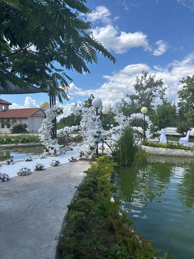An outdoor wedding ceremony setup is arranged next to a tranquil pond on a bright, cloudy day. The scene is located in the city of Panchevo, Bulgaria. A white carpet runs along a concrete path, flanked by small white floral arrangements. A decorative archway, also adorned with white flowers, stands over a small bridge crossing the pond. Two white chairs and a table covered with a white cloth are visible on the opposite bank of the pond. Green trees and a building with a red-tiled roof are in the background. The sky is a vibrant blue with scattered white clouds. The pond's surface is a rich green, reflecting the surrounding foliage and sky.