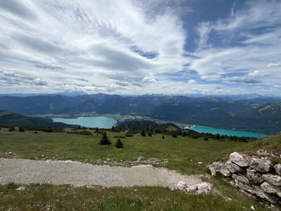The image captures a panoramic view from a mountain overlooking two turquoise lakes and a vast mountain range. In the foreground, a grassy slope descends towards a gravel path. Small evergreen trees are scattered across the slope. The middle ground features the lakes, nestled between rolling green hills and dense forests. Beyond the lakes, a chain of majestic mountains stretches across the horizon, their peaks partially obscured by clouds. The sky is a mix of blue and white, with wispy clouds creating a dynamic atmosphere. The scene evokes a sense of natural beauty and tranquility, suggesting the location is likely a popular hiking or scenic viewpoint in the Austrian Alps, possibly near Sankt Gilgen, known for its stunning lakes and mountainous terrain. There are no people or animals visible in the image.