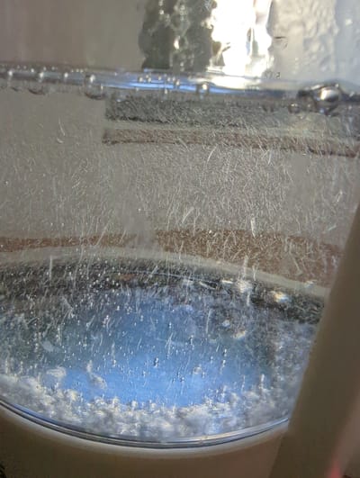 The image shows a close-up view of a clear glass container filled with a blue liquid and what appears to be effervescence or bubbles. The container's surface is covered in a fine network of white, thread-like structures, which might be ice crystals or mineral deposits. Horizontal bands of a brown, textured material are visible through the glass, possibly indicating the presence of a filter or another component within the container. The liquid itself has a clear blue hue and is agitated, with numerous small bubbles rising to the surface and some larger ones visible at the bottom. The lighting in the image suggests it is artificial and coming from above, creating reflections on the surface of the liquid and the glass. There is no indication of people or any specific location in the city of Taucha, Germany, within this image.