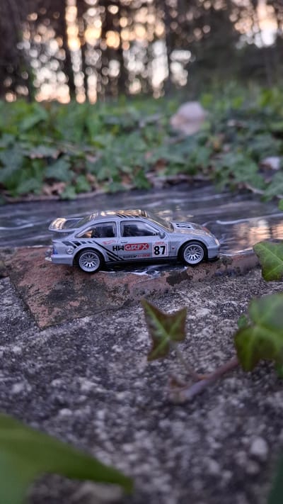 A close-up shot displays a silver toy car, a model race car with the number 87 on its side, positioned on a textured, likely brick or stone ledge covered in patches of green moss and climbing ivy. The background features blurred green foliage and a body of water with sunlight reflecting off its surface.
The image lacks sufficient information to pinpoint the exact location in Podbadac, Montenegro. There are no discernible identifying landmarks or features that enable precise geographical positioning.