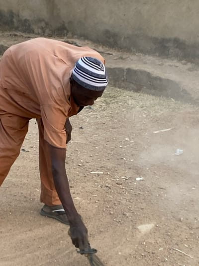 A person with dark skin, wearing a black and white striped cap and an orange outfit, stands on a dirt ground. The individual holds a bundle of sticks in their right hand. The ground is covered in dry vegetation. A concrete wall or structure is on the left side of the frame.