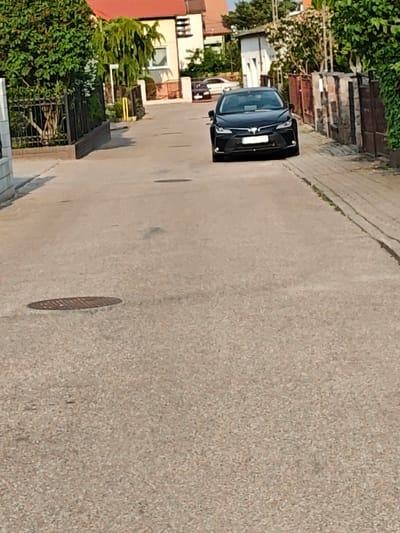 The image displays a residential street in Mława, Poland, under bright, clear daylight. A black 12th-generation Toyota Corolla sedan occupies the center of the frame, facing the viewer. The vehicle's front license plate is partially visible, consistent with a Polish registration. The road surface consists of light-colored, granular asphalt. A circular metal manhole cover is visible on the left side of the road, approximately halfway into the frame. Along the right side of the road, a concrete or paved sidewalk is present, bordered by a fence constructed from rough-hewn stone and dark wood panels. On the left side of the road, a black wrought-iron fence partially obscured by green vegetation separates the street from an adjacent property. In the background, multiple residential structures are visible, including a two-story building with a red-tiled roof and light-colored walls on the left, and a smaller white building further in the distance on the right. Abundant green trees and shrubs line both sides of the street, suggesting lush vegetation. No human figures are present in the scene.