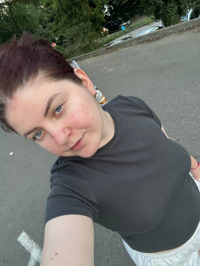The image is a selfie taken outdoors, likely in a parking lot or alongside a road. The subject is a woman with auburn hair pulled back, wearing a dark grey ribbed t-shirt and light-colored shorts. She has blue eyes and fair skin with some redness on her cheeks. In the background, trees and parked cars are visible. The ground is asphalt with white markings. The specific location in Lovran, Croatia, is not discernible from the visual information in the image.