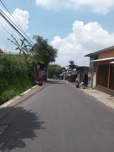 The image shows a street scene in what appears to be a village. The road is asphalt and runs straight into the distance. On the left side of the road, there is a wall and dense green vegetation. On the right, there are various small buildings with diverse architectural styles, including corrugated metal roofs and faded paint. Several motorcycles are parked along the side of the road, and in the distance, a few people and motorcycles are visible. Overhead, there are power lines. The sky is blue with scattered white clouds. There is no clear indication that this scene is Xayabury District, Laos.