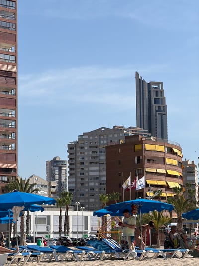 The image depicts a sun-drenched beach scene in Urbanització Cales I Talaies, Spain, with the azure sky serving as a backdrop. In the foreground, rows of empty blue lounge chairs and umbrellas are set up on the sand, awaiting beachgoers. Several people are visible in the mid-ground; one man stands near a lounge chair, looking at his phone, while a woman bends down. Further back, a couple sits in lounge chairs, and other individuals are scattered around. The background is dominated by a cluster of tall, modern buildings, characteristic of a coastal city. The architecture varies, from a curved brick building with yellow awnings to a sleek, grey skyscraper. Palm trees are interspersed among the buildings and on the beach, adding a tropical feel to the urban landscape. The overall impression is one of a vibrant tourist destination.