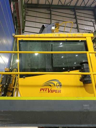 The image shows a close-up of the side of a yellow vehicle, possibly a piece of heavy machinery, with a dark gray lower section and a yellow railing in the foreground. The vehicle has large, multi-pane windows that appear to be covered in water droplets. A logo featuring a stylized snake head with the text "PIT VIPER SERIES" is visible on the yellow surface below the windows. To the left, there are mechanical components and equipment attached to the vehicle. The background appears to be an industrial building with a metal roof structure and blue siding. The scene is set indoors, likely in a workshop or hangar, and there are no people visible. The image was taken in Қопа ауылдық округі, Kazakhstan.
