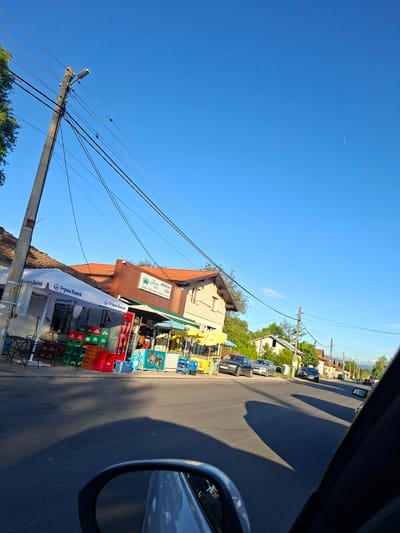Here is a description of the image:

The image shows a street scene, possibly in Drugan, Bulgaria. A two-story building with a red-tiled roof is on the left side of the street. The building has a sign that reads "Горна Баня" and appears to be a small shop selling groceries and drinks. There are beverage crates stacked along the sidewalk in front of the shop.

A utility pole is visible near the shop, with power lines running across the sky. The street is paved and leads into the distance. Several parked cars are visible on the opposite side of the street. The sky is clear and blue. The image is taken from the inside of a car, as part of the car's rearview mirror can be seen on the right side of the frame.