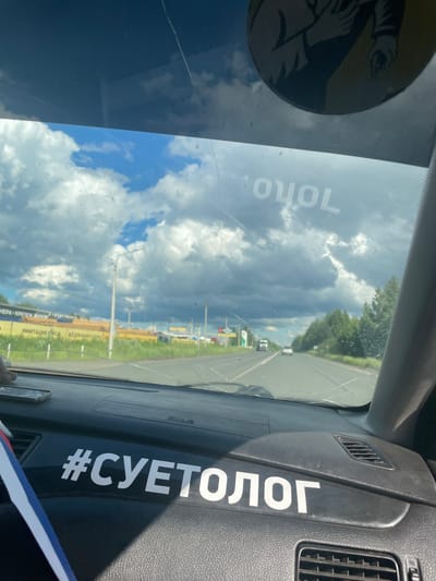 The image is a medium shot, taken from inside a vehicle looking forward through the windshield. The perspective is slightly angled, showing the dashboard on the right side of the frame and the windshield taking up most of the image. The sun is bright, indicating it is daytime, and there are scattered clouds in the sky, suggesting partly cloudy weather. The road ahead appears to be a highway with some traffic, surrounded by green foliage and trees.
On the dashboard, a white hashtag symbol followed by Russian text "#СУЕТОЛОГ" is prominently displayed. On the windshield, closer to the viewer and on the left side of the frame, another line of white Russian text reads "ТОЛО". A circular sticker, mostly in shadow, is also visible on the upper left of the windshield. The overall scene depicts a road trip or drive on a sunny day in what appears to be a rural or suburban setting. The cracked windshield adds a notable detail to the interior of the vehicle.