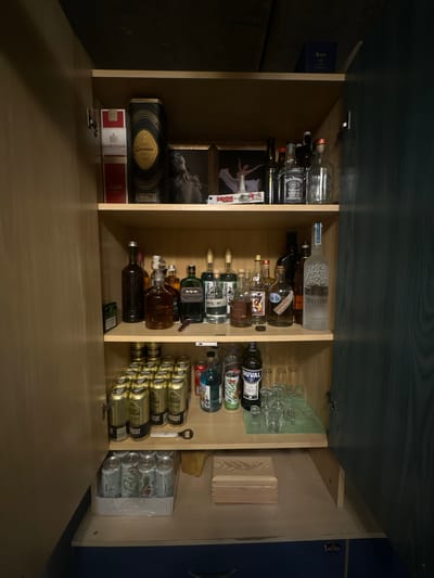The image shows an open wooden cabinet containing a collection of alcoholic beverages and related items. The cabinet is located inside a room with a dark, textured ceiling and dark wooden doors that are partially ajar.
The top shelf of the cabinet displays several bottles of liquor, including Courvoisier, Codorniu Cava, and Jack Daniel's. There are also two framed pictures on this shelf, one with a woman's silhouette and another with a figure in a white dress. A small blue box with text is visible on the top of the cabinet.
The second shelf is filled with a variety of liquor bottles, including those from brands like "3 LITTLE PIGS" and "BELVEDERE VODKA." Several smaller bottles and glasses are also present.
The third shelf holds rows of canned beverages, appearing to be beer, with labels such as "VALRAS BELMON" and "DRAUGHT." A can of Coca-Cola and a bottle of what appears to be blue-colored liquor are also on this shelf. Some glassware is arranged on a green mat.
The bottom shelf has a box of canned beverages, possibly more beer, and a small wooden box with an engraved logo. A bottle opener lies on the shelf above.
The environment suggests a home bar or storage area. The specific location in Lapės, Lithuania, is not directly indicated by any visual cues within the image itself.