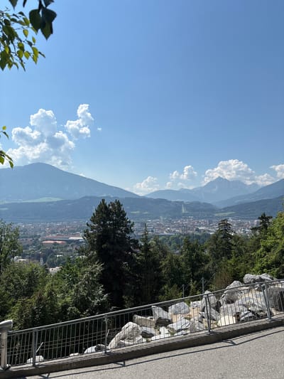 This is a vertical, wide-angle, outdoor photograph captured on a sunny day. The scene overlooks a city nestled in a valley between towering mountains, characteristic of Innsbruck, Austria. The sky is a clear, bright blue, with a few scattered, fluffy white clouds adding texture.

In the foreground, to the right, a metal railing runs along a path made of asphalt, casting a distinct shadow. Behind the railing, a rocky embankment is visible, filled with large, grey stones of various sizes. Lush green trees and foliage line the left side of the embankment and spill into the midground.

The midground is dominated by the city, with its densely packed buildings featuring red-tiled roofs. A church spire can be seen poking above the rooftops. Beyond the city, the majestic mountains rise, their slopes covered in a mix of dark green forests and lighter green patches. The mountain ranges fade into a hazy blue in the distance, suggesting depth and scale.

The lighting indicates it is likely daytime, possibly late morning or early afternoon, given the bright sunlight and clear skies. There is no text visible in the image. The overall mood is serene and picturesque, showcasing the beautiful natural landscape of the Alps surrounding the urban environment.