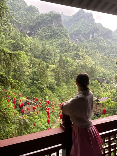 A female person with brown hair tied back is seen from behind, leaning over a wooden railing on a balcony, viewing a landscape of mountains and greenery. The person is wearing a white, long-sleeved shirt and a pink skirt. The balcony structure is made of dark wood. In the background, there are green mountains with some visible rock faces. A dense forest of trees covers the lower areas, interspersed with red lanterns. There is a small wooden structure with a peaked roof, suspended amidst the trees and lanterns. The location is most likely in Yongding District, China. The sky is overcast and the light is diffused, suggesting a misty or cloudy day.