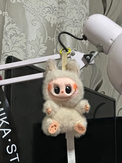 This media file features a close-up shot of a white, fluffy toy figure with large, cartoonish eyes and pink rosy cheeks. The toy is lying on its back, with its limbs spread out. It appears to be attached to a white, adjustable lamp or stand by a yellow loop. A black wire extends from the lamp.
In the background, there is a textured, light-colored surface with a floral pattern, possibly wallpaper or fabric. Behind the toy and lamp, a dark, flat surface is visible, which could be a desk or electronic device. A black ribbon with white text is partially visible in the lower right corner.
The lighting suggests an indoor setting, with no indication of time of day or weather. The focus is on the toy and the immediate surroundings, creating a sense of playful intimacy. No specific emotions are conveyed by the inanimate subjects.
Visible text on the ribbon appears to read "MIKA•ST", though it is partially obscured. A small white tag is also attached to the toy's leg, bearing some symbols and text that are too small to decipher. The overall impression is of a display or arrangement of decorative items. The location context is Bishkek, Kyrgyzstan.