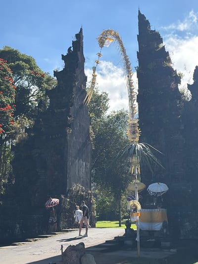 A man and a woman stand on an asphalt path near a stone structure. The man wears a dark t-shirt and shorts, while the woman wears a white dress. To their left, a red and white checkered umbrella is propped up. Ahead of them, two tall, dark stone gate-like structures flank a path leading through an archway decorated with a long, hanging floral arrangement. A small altar with offerings and umbrellas is visible to the right of the archway. Lush green trees and a bright blue sky with scattered clouds form the background. The scene is illuminated by sunlight. This image appears to be taken at the Handara Gate in Sukasada, Indonesia.