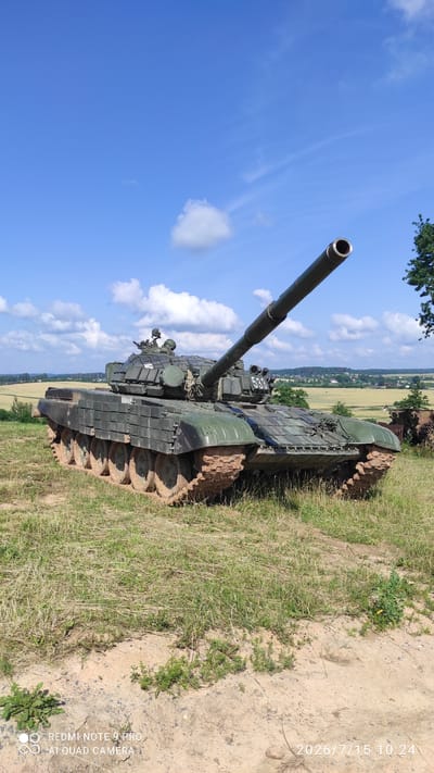 An outdoor, eye-level, medium shot captures a dark green Soviet-era T-72 tank positioned on a grassy, dirt embankment under a bright blue sky with scattered white clouds. The tank is angled slightly to the right, with its cannon barrel extending upwards and to the right. Mud is caked on the tank's tracks and lower hull. In the background, a rural landscape unfolds, featuring green fields, scattered trees, and a village with buildings visible in the distance. The ground in the foreground is covered in dry grass and dirt. The bottom right corner of the image displays a timestamp and device information: "2026/7/15 10:24" and "REDMI NOTE 9 PRO AI QUAD CAMERA".